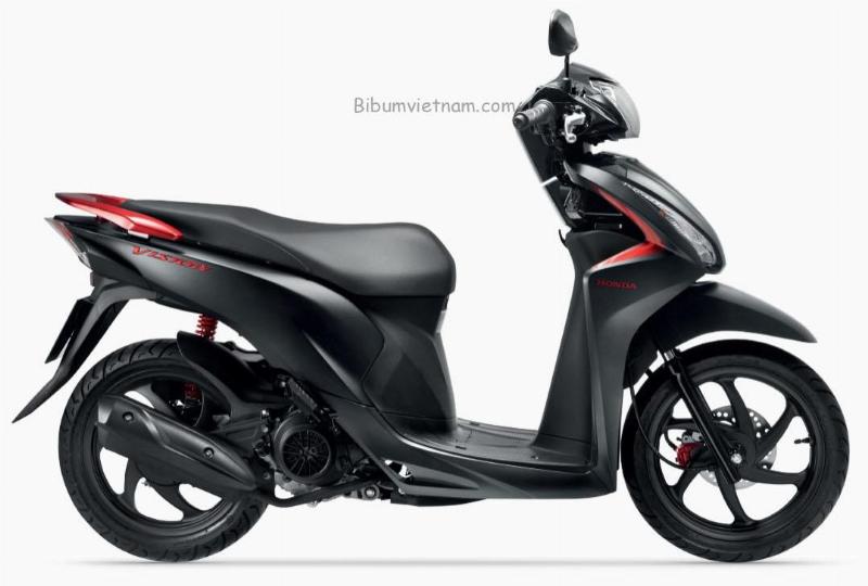 Một chiếc xe Honda Wave Alpha 110 màu đỏ đang di chuyển trên con đường đông đúc ở Việt Nam, phía sau là những tòa nhà và xe cộ khác.