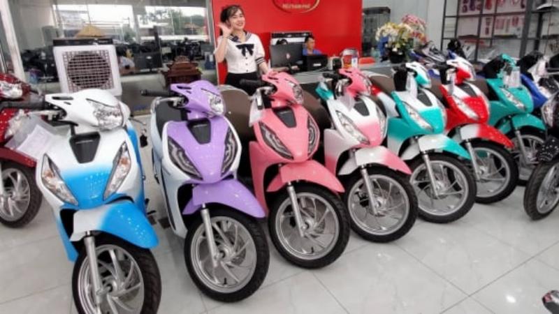 Một chiếc xe Honda Vision màu trắng đen đang di chuyển trên một con đường có cây xanh ở khu vực đô thị Việt Nam. Hình ảnh thể hiện Vision là phương tiện di chuyển phổ biến và phù hợp với cảnh quan đô thị.